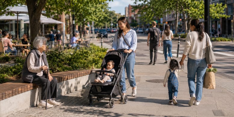 Woman pushing a baby stroller near an older woman seated on a bench, while another woman walks with a child along a tree-lined street.