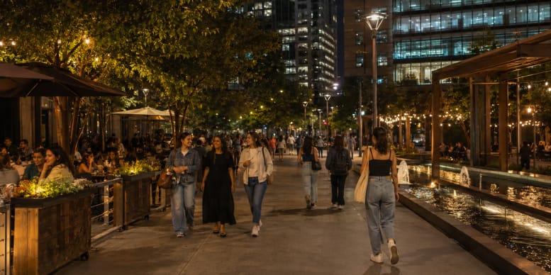 Women walking through a busy urban public space at night with restaurants, trees, lighting and a water feature.